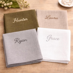 May include: Four square fabric napkins folded neatly, each featuring a different embroidered name in cursive script. The colours include olive green with the name Hunter, beige with Laura, light grey with Ryan, and white with the name Grace.