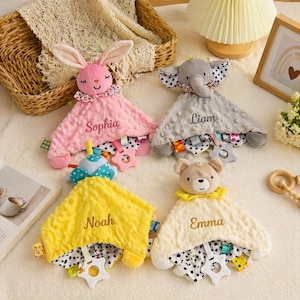 May include: Four plush baby security blankets with animal heads: a pink bunny, a gray elephant, a yellow duck, and a cream-colored bear. Each has a name embroidered on it: Sophia, Liam, Noah, and Emma. The blankets have a patterned border.