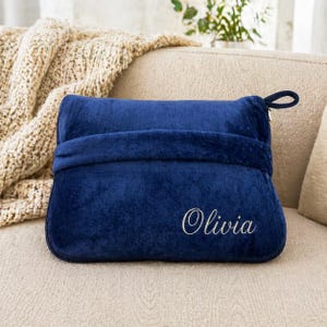 Puede incluir: Una almohada de felpa azul marino con el nombre "Olivia" bordado en escritura blanca. La almohada tiene un asa de bucle y una banda envolvente. Una manta beige es visible en el fondo.