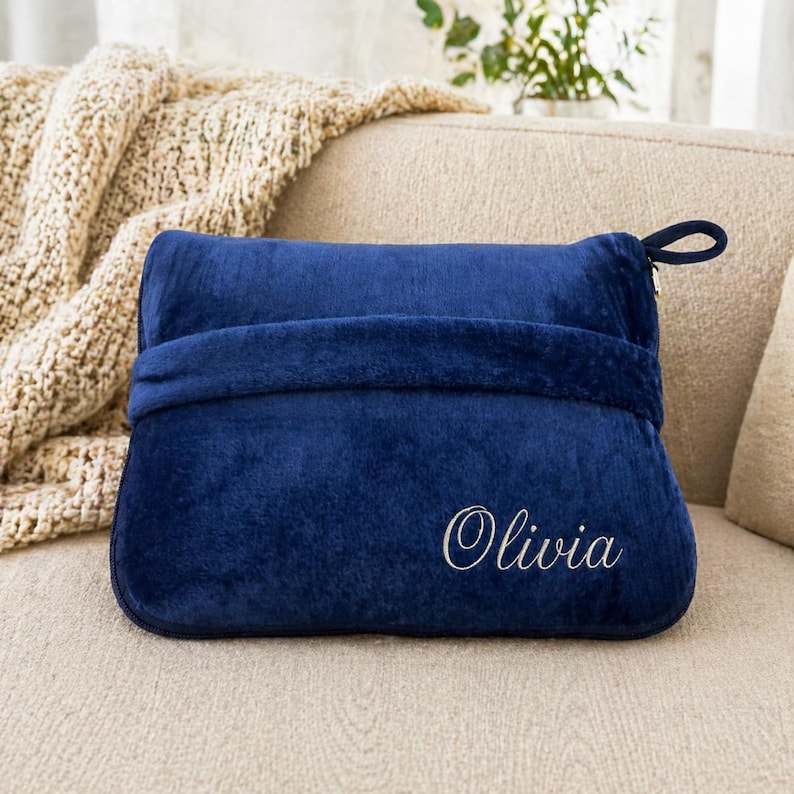 Puede incluir: Una almohada de felpa azul marino con el nombre "Olivia" bordado en escritura blanca. La almohada tiene un asa de bucle y una banda envolvente. Una manta beige es visible en el fondo.
