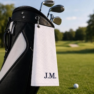 Op de afbeelding: Een wit golfhanddoekje met wafelstructuur en de in marineblauw geborduurde initialen J.M., hangend aan een zwart-witte golfbag. De handdoek is voorzien van een metalen oogje en clipbevestiging en heeft een textuurpatroon over het gehele oppervlak.