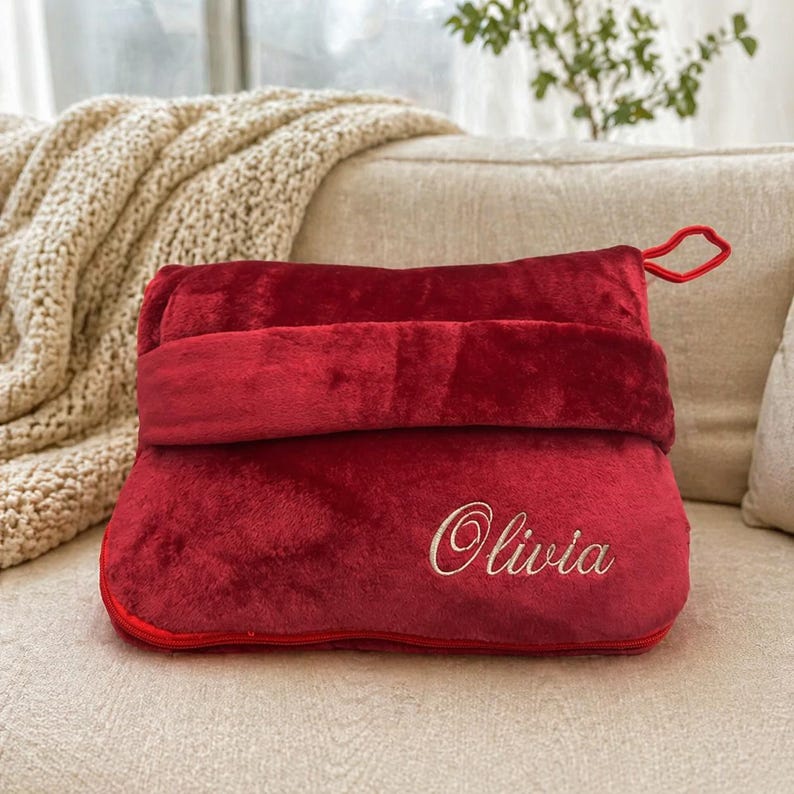 Puede incluir: Una almohada de viaje roja y afelpada con un asa de bucle. La almohada tiene un dise&ntilde;o plegable y est&aacute; bordada con el nombre "Olivia" en escritura blanca. La almohada est&aacute; sobre un sof&aacute; beige con una manta color crema.