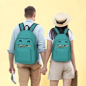 Puede incluir: Dos mochilas de color verde azulado con una cara de dibujos animados con los ojos cerrados, expresión de aburrimiento y una boca pequeña. Cada mochila tiene correas negras, un compartimento principal y un bolsillo frontal. El diseño es idéntico.