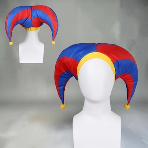 Può includere: Un cappello da giullare con pannelli in tessuto rosso e blu alternati, una fascia gialla e piccoli pon pon gialli alle estremità delle due punte. Il cappello è mostrato su una testa di manichino bianca, con vista frontale e posteriore.