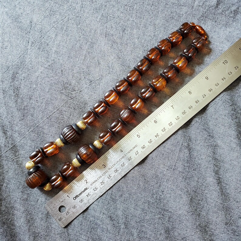 May include: A vintage amber-colored beaded necklace with a variety of bead shapes, including round and barrel-shaped beads. The necklace is approximately 28 inches long, with gold-colored spacers and dark brown accents. A ruler is placed next to the necklace.