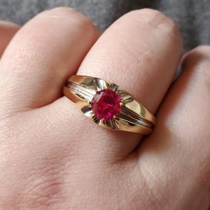 May include: A gold ring with a large, oval, red gemstone at the center. The ring has a decorative, ridged design on the sides and a floral-like setting around the gemstone. The ring is on a finger.