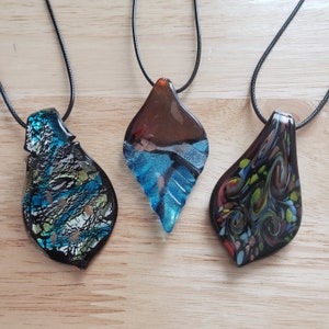 May include: Three glass pendants on black cords. The pendants are shaped like teardrops and feature abstract designs in shades of blue, brown, black, green, and gold.