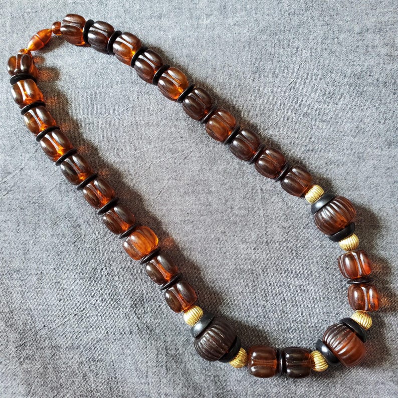May include: A necklace featuring amber-colored, barrel-shaped beads with black spacers. The necklace has a mix of smooth and ribbed beads, with gold-colored accents. The beads are strung together, creating a unique jewelry piece.