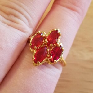 May include: A gold-toned ring featuring a cluster of five marquise-cut, vibrant red gemstones. The gemstones are arranged in a floral pattern, with a textured gold band. The ring is shown on a finger.
