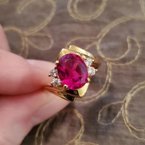 May include: A gold ring featuring a large, oval-cut, magenta gemstone as the centerpiece. The ring is accented with smaller, round, clear gemstones. The ring's band and setting are a polished gold color.