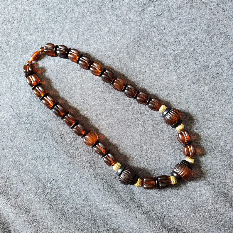May include: A necklace featuring amber-colored, barrel-shaped beads. The beads are strung together with smaller, darker brown beads and gold-colored spacers. The necklace is displayed on a gray surface.