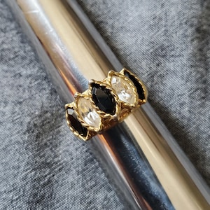 May include: A gold-toned ring featuring alternating black and clear marquise-cut stones. The ring is set on a silver metal object against a gray fabric background. The ring's design includes a twisted rope-like border around each stone.