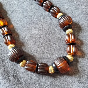 May include: A necklace featuring a variety of brown and amber-colored beads. The beads are interspersed with black and gold-colored accents. The necklace is displayed on a gray surface.