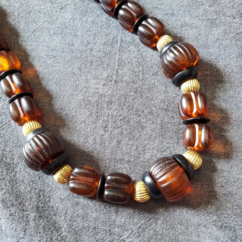 May include: A necklace featuring a variety of brown and amber-colored beads. The beads are interspersed with black and gold-colored accents. The necklace is displayed on a gray surface.