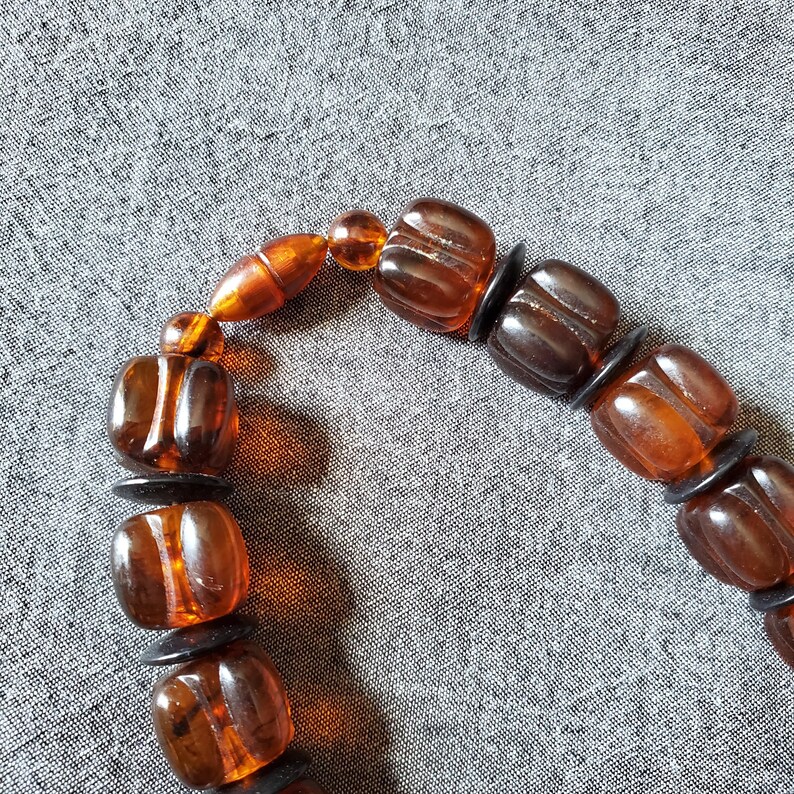 May include: A close-up of a bracelet featuring amber-colored, square beads with rounded edges. The beads are separated by small, black, circular spacers. The bracelet has a warm, earthy tone, with the beads appearing translucent.