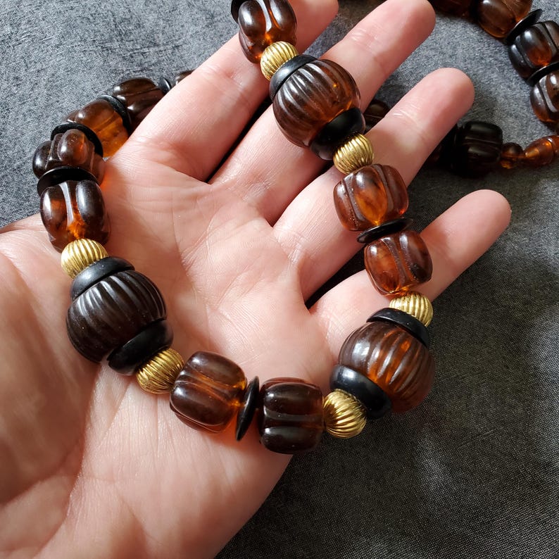 May include: A vintage necklace featuring large, amber-colored beads with ribbed detailing. The beads are interspersed with black and gold-toned accents, creating a striking contrast. The necklace is displayed against a neutral background.
