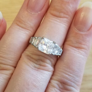 May include: A silver ring with a large square-shaped clear cubic zirconia stone surrounded by smaller clear cubic zirconia stones.