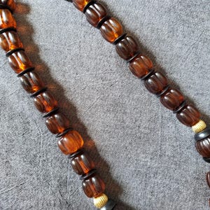 May include: A close-up of a necklace featuring amber-colored, barrel-shaped beads. The beads are separated by small black spacers. The necklace has a gold-colored accent near the clasp and is set against a gray background.
