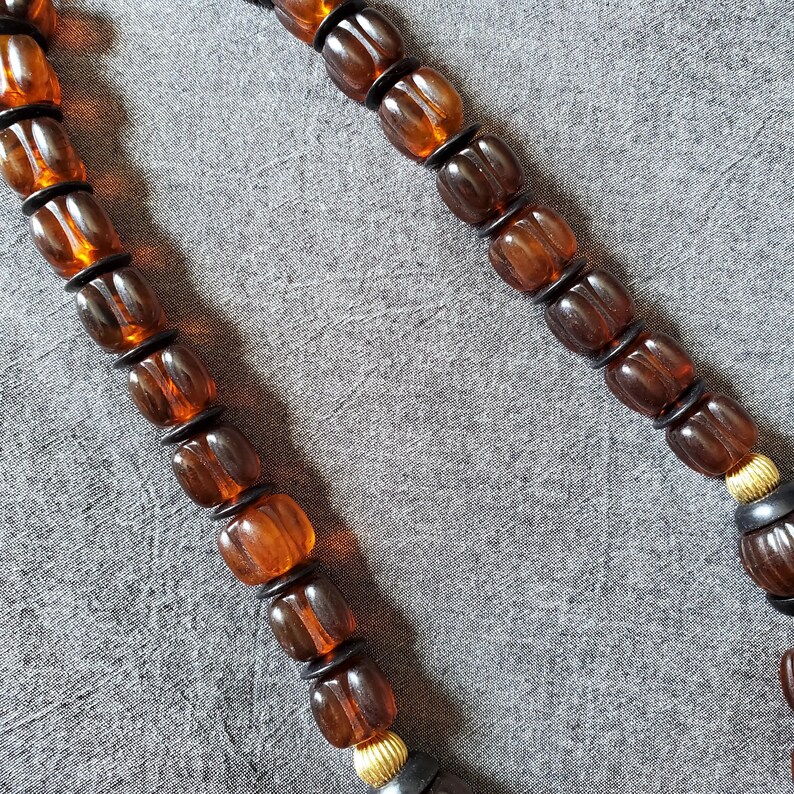 May include: A close-up of a necklace featuring amber-colored, barrel-shaped beads. The beads are separated by small black spacers. The necklace has a gold-colored accent near the clasp and is set against a gray background.