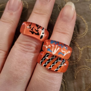 May include: Three orange glass rings with abstract designs in black, white, and blue. The rings are stacked on a finger.