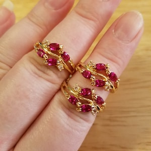 May include: Gold-toned rings with a leaf-like design, featuring oval-shaped, ruby-red gemstones and small, clear accent stones. The rings are displayed on a light-colored surface, showcasing their intricate details and vibrant color.