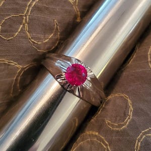 May include: A silver ring with a vibrant, round, red gemstone at the center. The gemstone is surrounded by a sunburst design. The ring is displayed on a shiny, silver cylindrical object, with a brown, patterned fabric in the background.