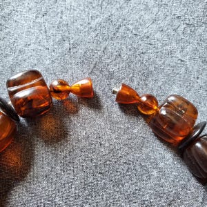 May include: Close-up of a necklace with large, amber-colored beads. The beads are a mix of rounded and rectangular shapes, with a visible clasp. The necklace is on a gray fabric background.