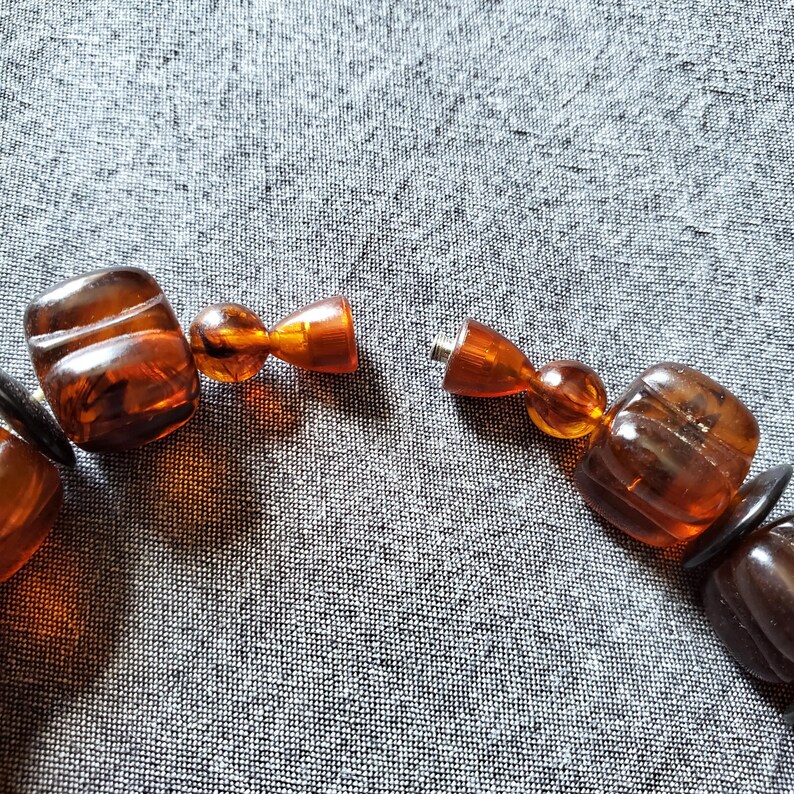 May include: Close-up of a necklace with large, amber-colored beads. The beads are a mix of rounded and rectangular shapes, with a visible clasp. The necklace is on a gray fabric background.