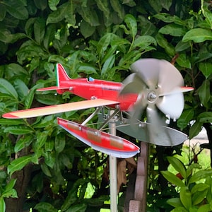 Pode incluir: Um cata-vento de hidroavião vermelho e creme com uma hélice giratória. O avião tem um corpo vermelho, asas creme e uma cauda vermelha e verde. O cata-vento é montado em um poste de metal e em um fundo de folhagem verde.