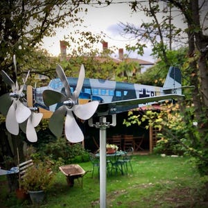 Op de afbeelding: Een metalen vliegtuigwindmolen met een camouflage-ontwerp, met meerdere propellers. Het model is op een paal in een tuin geplaatst, met bomen en een huis op de achtergrond. Het vliegtuig heeft de letters "DT" op de vleugel.