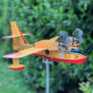 Può includere: Un modello di aereo giallo e rosso con due eliche e il numero 21 sulla parte anteriore. L'aereo ha la scritta "VIGILI DEL FUOCO" e la bandiera italiana sulla coda. Le ali sono rosse e gialle.