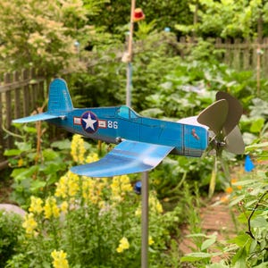 Peut inclure: Un moulin à vent en forme d'avion bleu en métal avec une hélice et le numéro 86 sur le côté. L'avion a un emblème d'étoile et est installé dans un jardin avec des fleurs jaunes et du feuillage vert.