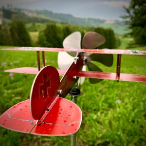 Pode incluir: Um moinho de vento biplano vermelho com uma hélice branca e uma barbatana traseira vermelha com um desenho de cruz branca. O avião de metal é colocado contra um fundo de relva verde e árvores.