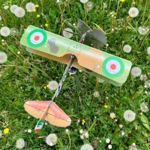 Può includere: Un aereo giocattolo con una combinazione di colori verde, arancione e bianco, con elica e coccarde. Le ali presentano un motivo mimetico, su uno sfondo di erba verde e denti di leone.