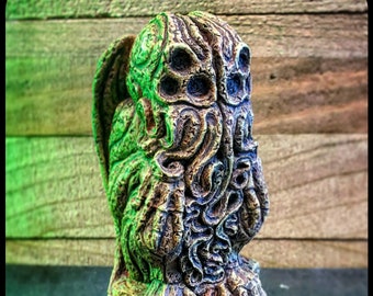 May include: A detailed, green, brown, and blue stone sculpture of Cthulhu, a fictional cosmic entity from the works of H.P. Lovecraft. The sculpture is in a seated position with its arms resting on a square base.