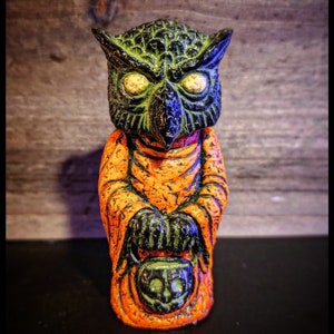 May include: A green and orange owl figurine with yellow eyes holding a black and green jack-o'-lantern. The owl is wearing a robe and has a textured surface.