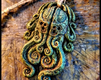 May include: A green and gold ceramic pendant with a Cthulhu design. The pendant is shaped like a head with tentacles and has a distressed finish. The text "CULT OF CTHULHU AMULET" is printed below the pendant.