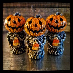 Può includere: Tre figurine in ceramica a tema Halloween con teste di zucca e mais dolce nelle mani. Le figurine sono dipinte in tonalità di arancione, nero e bianco.