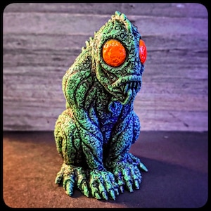 May include: Green and blue textured figurine of a creature with large orange eyes and a toothy grin. The creature is sitting with its legs crossed and its arms resting on its knees.