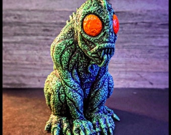 May include: Green and blue textured figurine of a creature with large orange eyes and a toothy grin. The creature is sitting with its legs crossed and its arms resting on its knees.