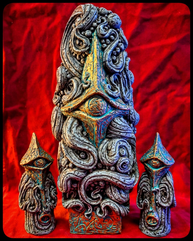 May include: Three gray, textured, ceramic sculptures with gold and blue accents. The tallest sculpture is a tall, thin, rectangular shape with a face-like design. The two smaller sculptures are shorter and wider, with similar face-like designs.