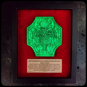 Puede incluir: Un artefacto verde fluorescente enmarcado que representa una criatura con una etiqueta de texto que dice "Yuggoth Artifact". La etiqueta describe el artefacto como una piedra tallada en una piedra luminosa no identificada que representa un "Mi-Go" o un "Old One".