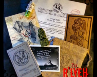 May include: A collection of items related to the fictional city of R'lyeh from the works of H.P. Lovecraft. The collection includes a small brown leather-bound journal, a small vial with green beads, a small green tassel, a postcard with a black and white photo of a rocky island, a brown paper bag with the text "The R'lyeh Collection" in red, and a brown card with a black and white illustration of a creature with wings.