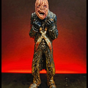 May include: A detailed figurine of a grotesque figure with a textured, peeling face and tattered clothing. The figure has a menacing expression and is posed with its arms crossed. The color palette includes shades of brown, black, and red, creating a spooky aesthetic.