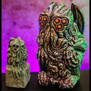 May include: Two green and brown Cthulhu figurines. The larger figurine has a wing and multiple eyes. The smaller figurine is standing on a square base.