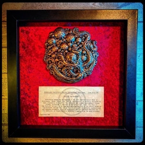 Puede incluir: Una vitrina enmarcada que muestra un objeto tridimensional de color dorado que se asemeja a un grupo de burbujas u orbes conectados por zarcillos. El objeto está etiquetado como "The Key of the Sothoth" y va acompañado de una descripción de texto sobre sus orígenes y significado.