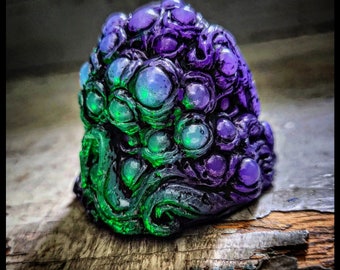May include: A small, hand-painted sculpture of a creature with a textured, bumpy surface. The creature is a dark purple and green color with many small, round eyes.