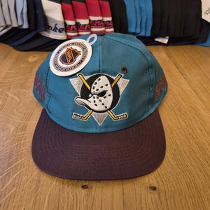 Op de afbeelding: Teal en paarse baseballcap met het Anaheim Ducks-logo, een hockeymasker en gekruiste hockeysticks. De cap heeft een paarse klep en een label met het NHL-logo. Op de achtergrond is een verzameling andere baseballcaps te zien.