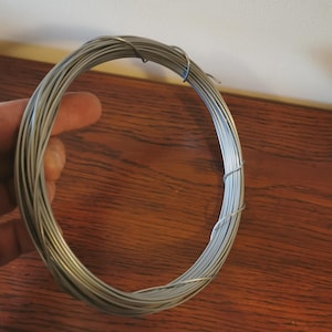 May include: A coil of silver-colored wire, tightly wound into a circular shape. The wire appears to be made of metal and is held against a wooden surface. The wire is likely used for crafting or other DIY projects.