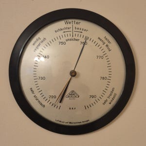 Könnte beinhalten: Ein Vintage-Barometer mit einem schwarzen kreisförmigen Rahmen. Das Zifferblatt zeigt eine Skala von 720 bis 790, mit Text, der Wetterbedingungen wie "windig" und "schön" anzeigt. Der Zeiger zeigt auf den Bereich "unsicher". Der Text "GOERZ BERLIN" ist sichtbar.